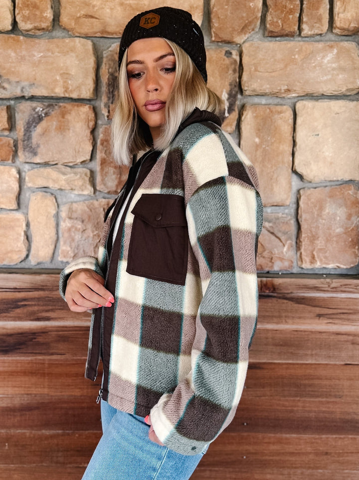 A woman wearing a brown, cream, and sage green plaid fleece jacket with a black beanie, posing in front of a rustic stone wall.