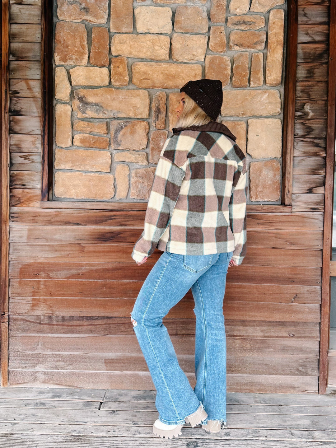 A woman stands on a wooden porch in front of a stone wall, wearing a brown, cream, and black buffalo plaid flannel shirt, blue jeans, and a black beanie.