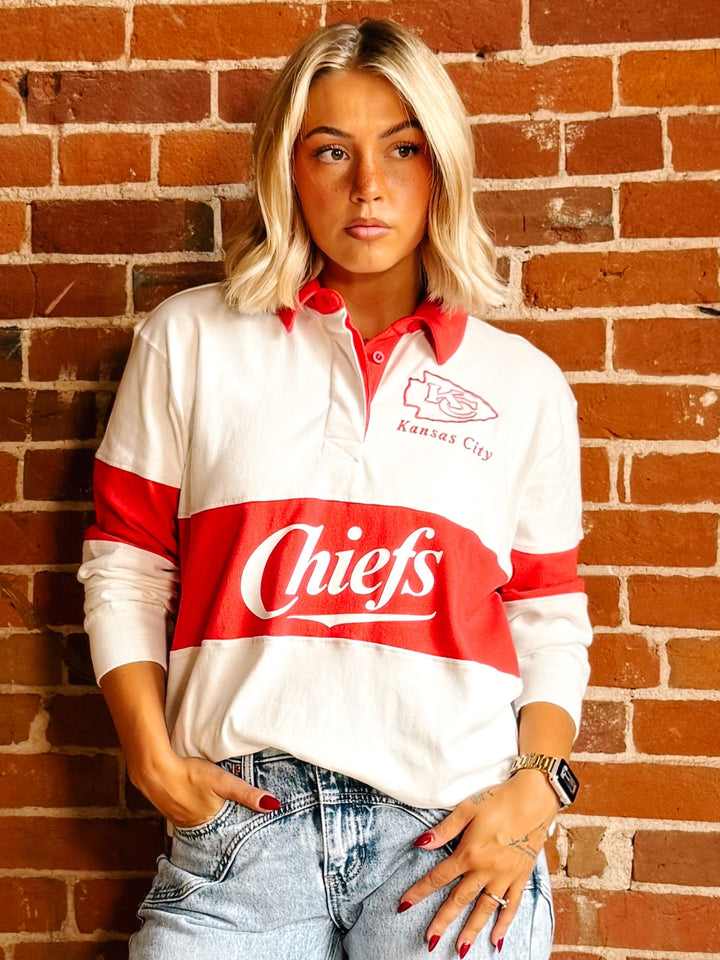 A woman wearing a Kansas City Chiefs long-sleeve rugby-style shirt with a red and white color-blocked design, standing against a red-brown brick wall.