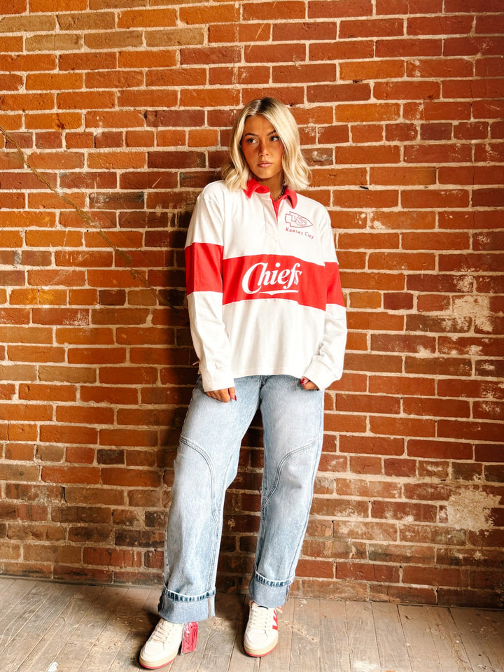A woman stands against a red-brown brick wall wearing a vintage white and red Coca-Cola Chief long-sleeve rugby shirt, light-wash blue jeans, and white sneakers.