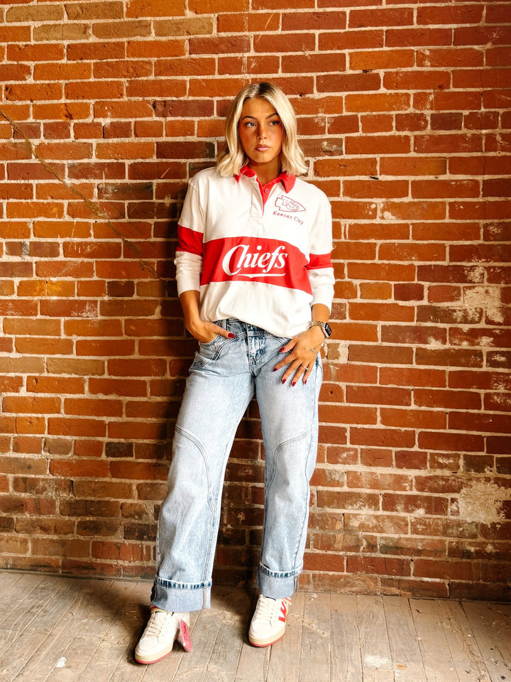 A woman stands against a red brick wall wearing a white long-sleeve polo shirt with a bold red horizontal stripe and &