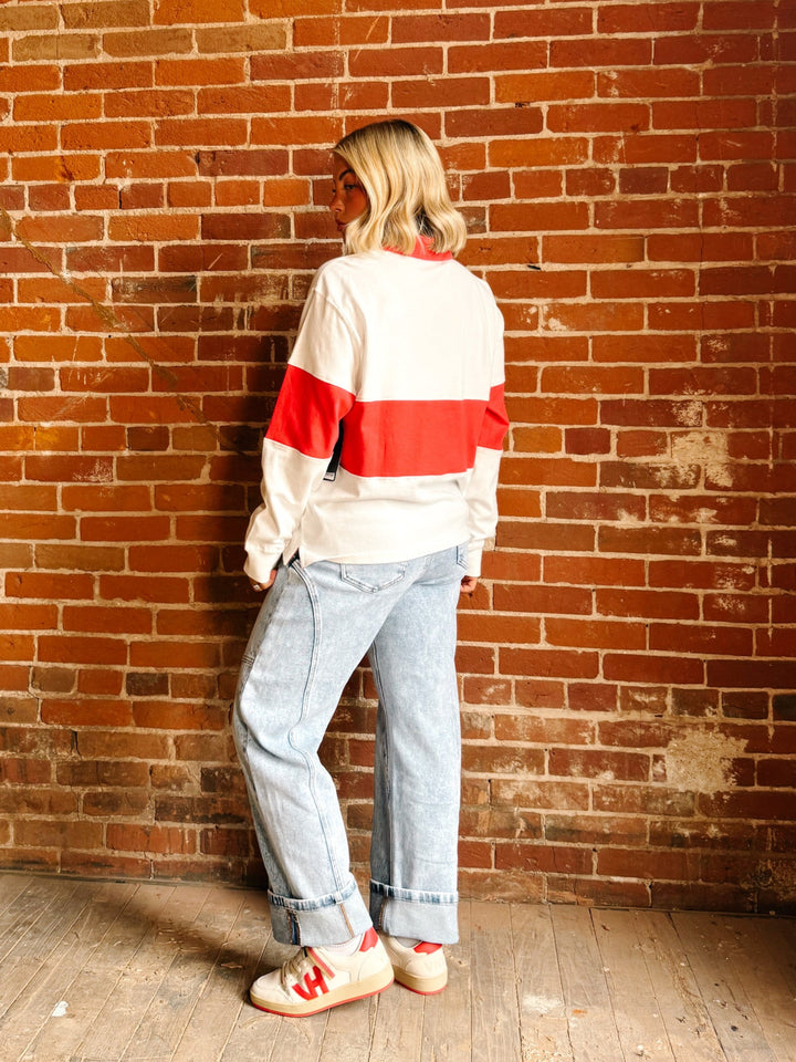 A woman stands with her back to the camera against a red-brown brick wall, wearing a white and red color-blocked sweatshirt, light-wash blue jeans, and white sneakers with red accents.