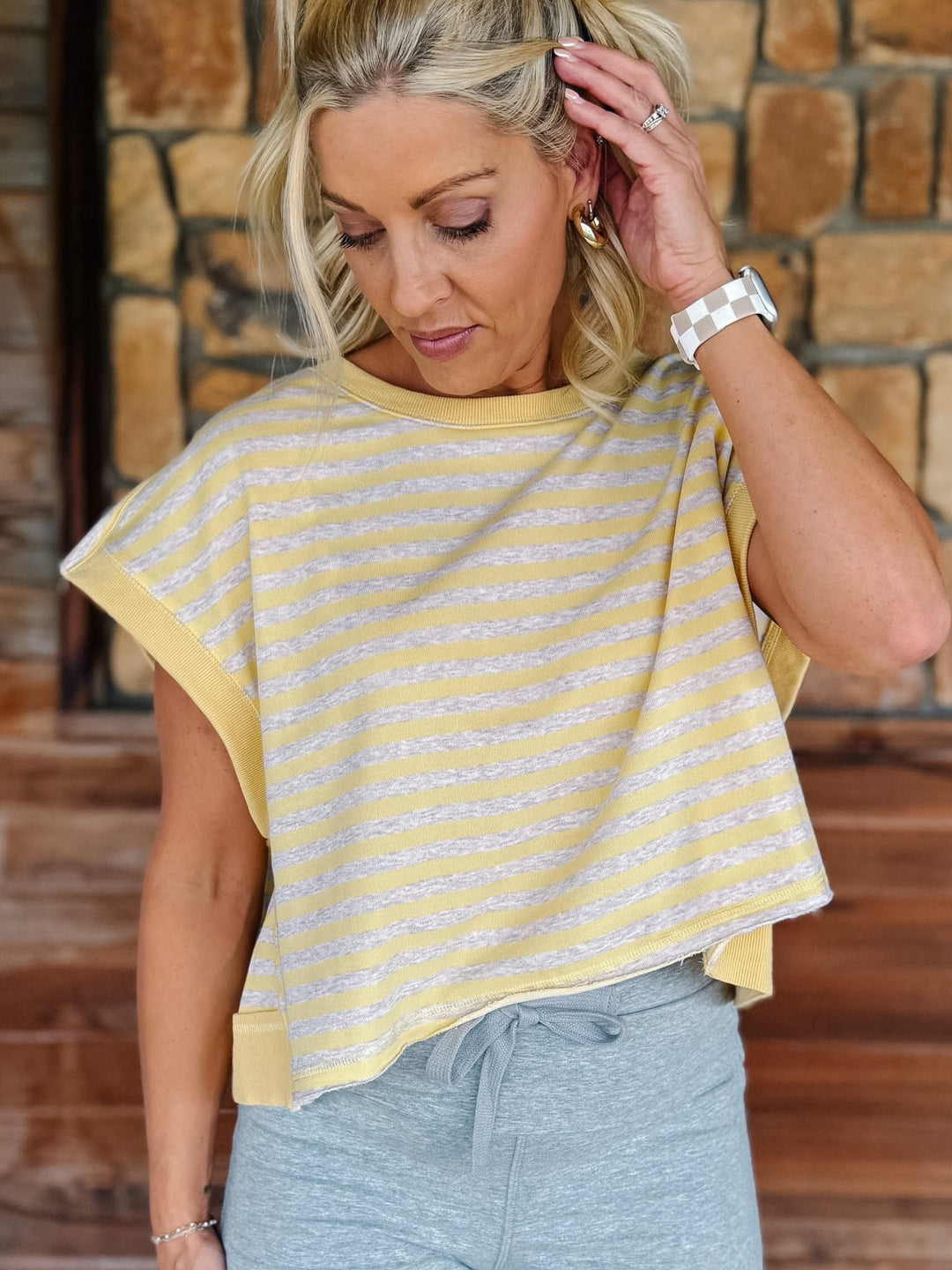 A woman wearing a yellow and heather oatmeal striped short-sleeve  top with gray drawstring sweatpants, posing in front of a rustic stone wall.