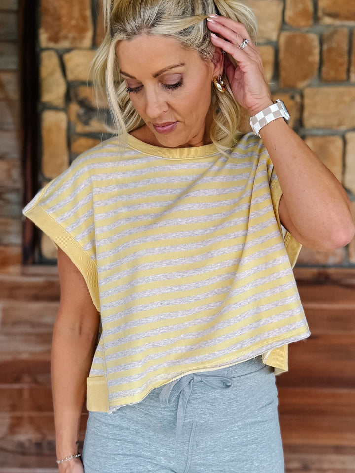 A woman wearing a yellow and heather oatmeal striped short-sleeve  top with gray drawstring sweatpants, posing in front of a rustic stone wall.