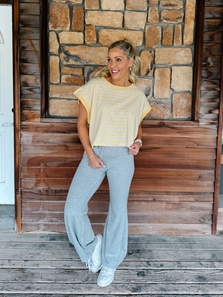 A woman wearing a yellow and heather oatmeal striped short-sleeve  top with gray drawstring sweatpants, posing in front of a rustic stone wall.