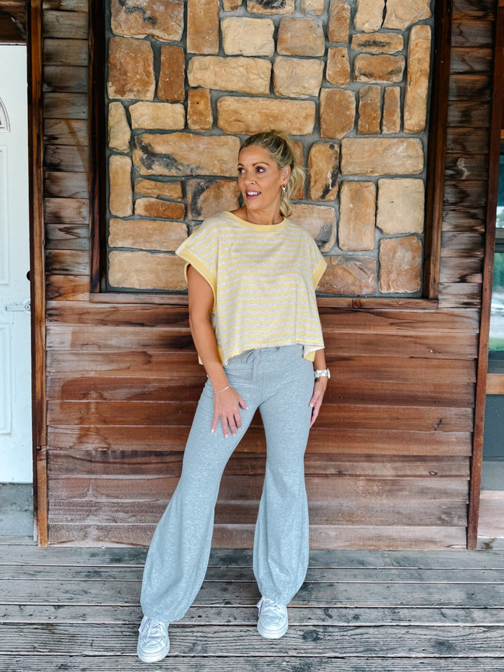 A woman wearing a yellow and heather oatmeal striped short-sleeve  top with gray drawstring sweatpants, posing in front of a rustic stone wall.