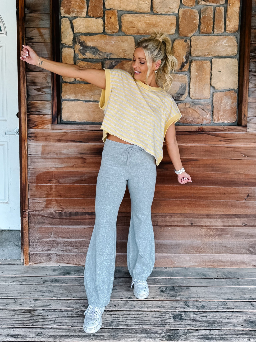 A woman wearing a yellow and heather oatmeal striped short-sleeve  top with gray drawstring sweatpants, posing in front of a rustic stone wall.