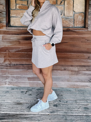 A woman wearing a heather taupe pullover and matching drawstring mini skirt, paired with light blue sneakers, standing on a rustic wooden porch with a stone wall background.