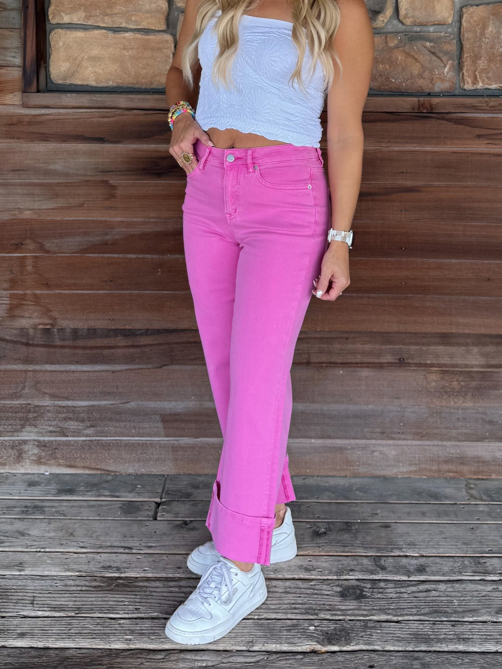 A woman wearing bright pink rolled-hem jeans, a white ribbed tank top, and white sneakers, standing on a rustic wooden deck in front of a stone wall.
