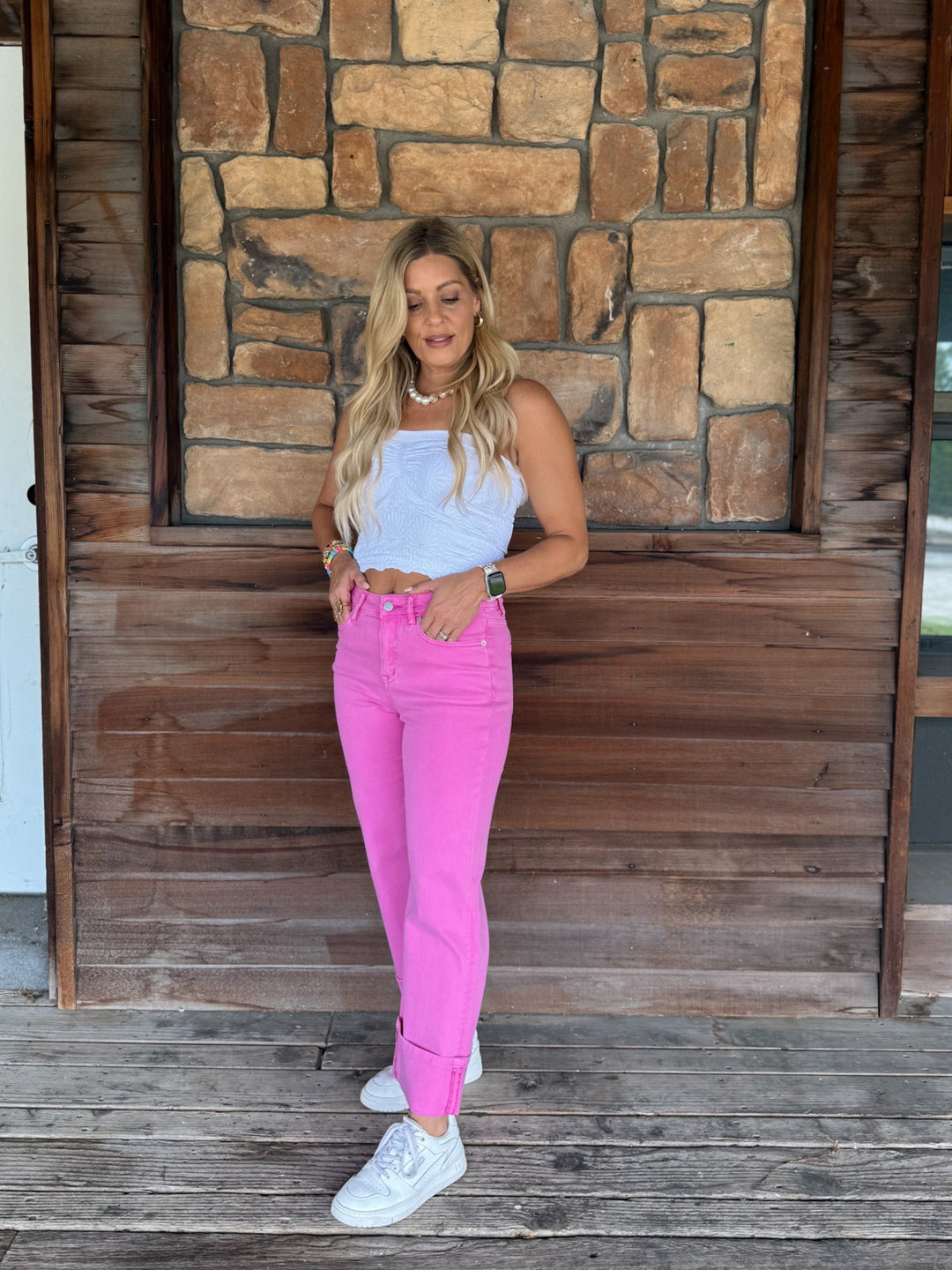 A woman stands on a wooden deck in front of a rustic stone and wood wall, wearing a light blue sleeveless top, bright pink skinny jeans, and white sneakers.