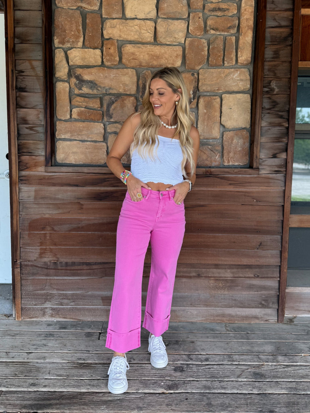 A woman modeling bright pink wide-leg jeans with a white crop top and white sneakers, standing on a wooden porch in front of a stone wall.