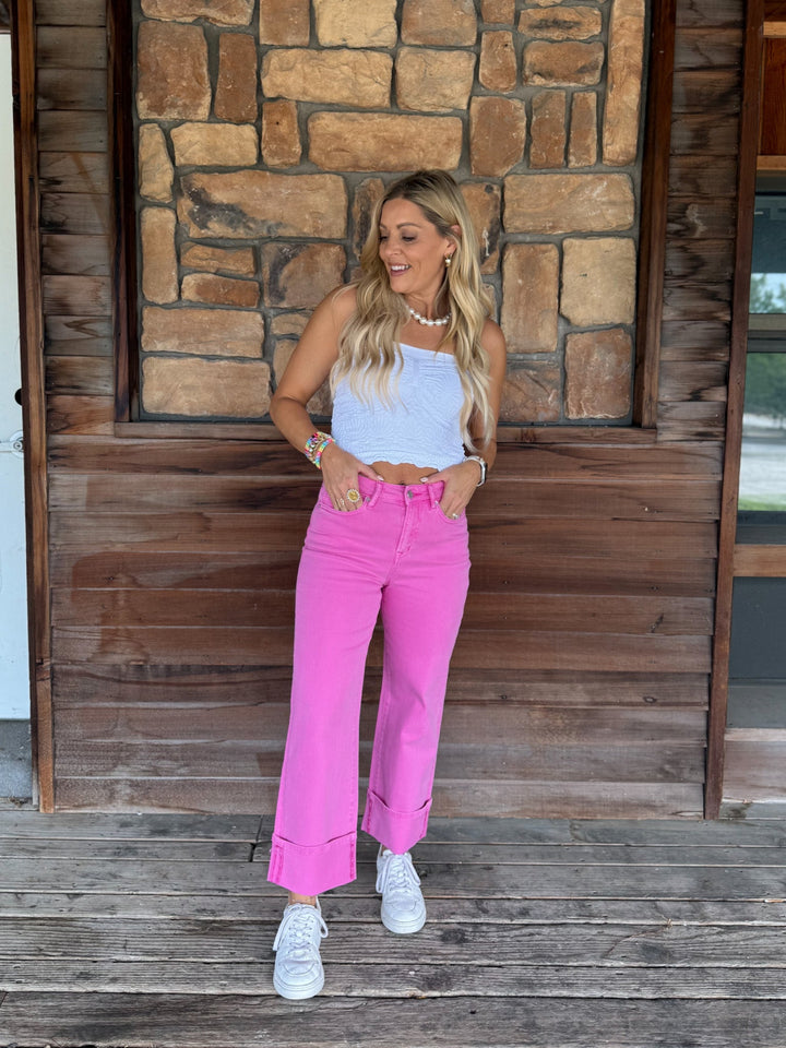 A woman modeling bright pink wide-leg jeans with a white crop top and white sneakers, standing on a wooden porch in front of a stone wall.
