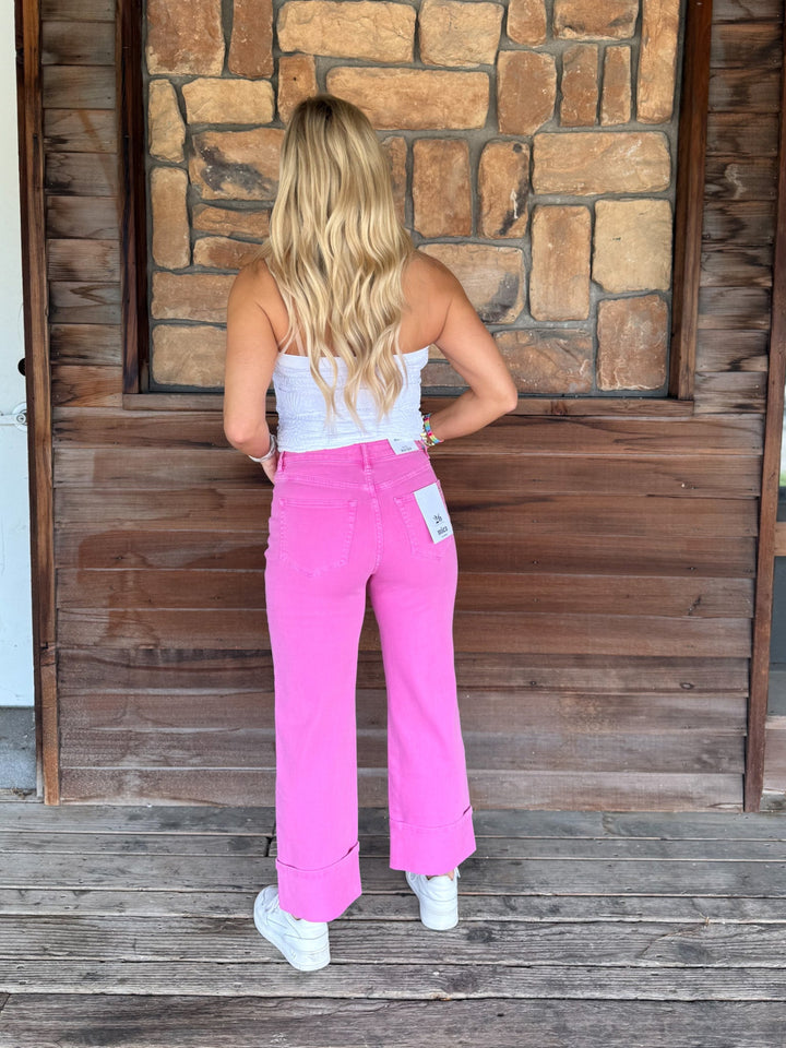 A woman stands with her back to the camera wearing bright pink wide-leg cropped pants, a white strapless top, and white sneakers, posed in front of a rustic wooden and stone wall.