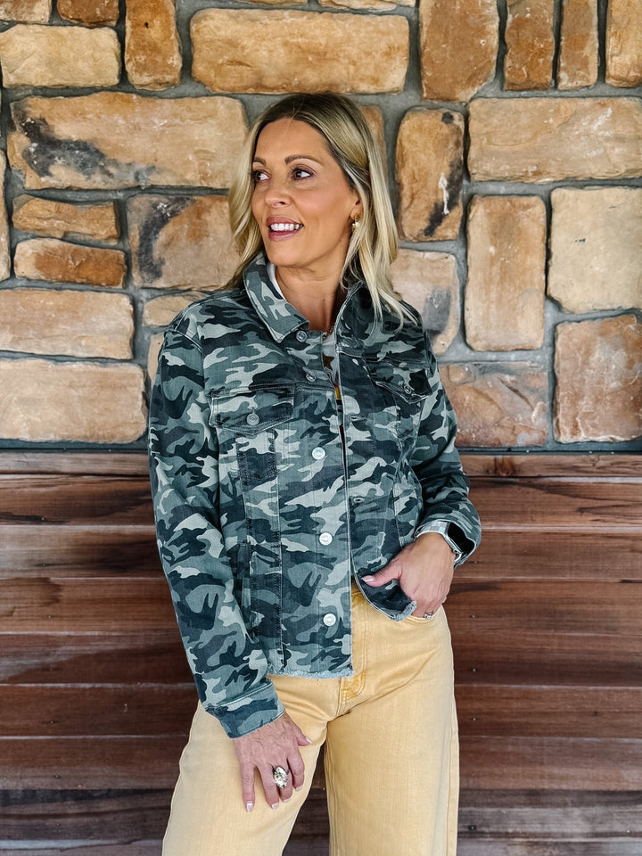A woman wearing a dark green and gray camouflage jacket with tan pants, standing in front of a rustic stone and wood wall.