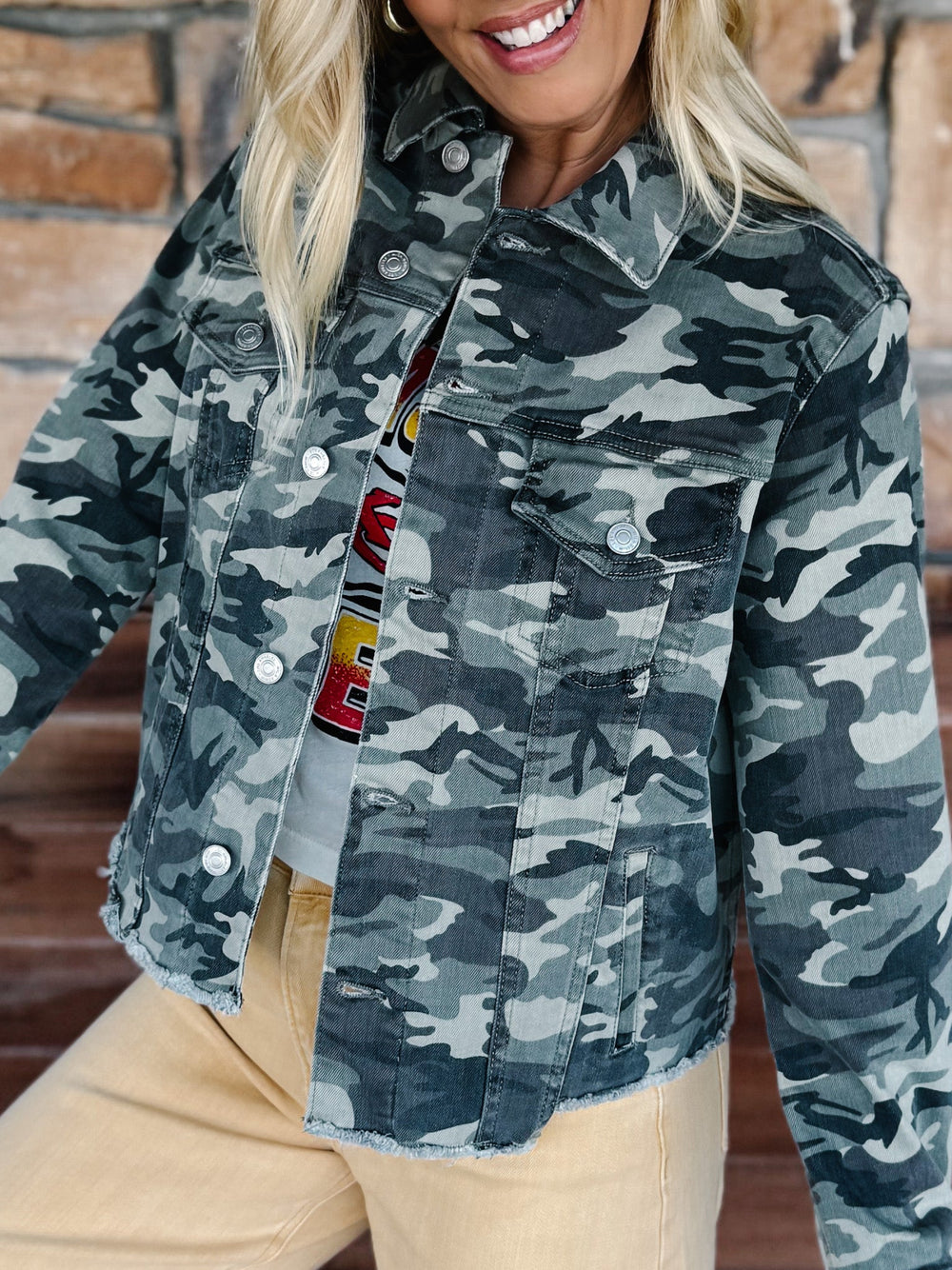A woman wearing a cropped camouflage denim jacket with a button-front closure and chest flap pockets, paired with beige pants, shown against a brick wall background.