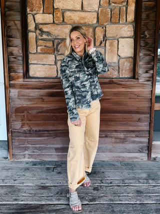 A woman stands on a rustic wooden porch in front of a stone wall, wearing a camouflage quarter-zip pullover jacket and light tan wide-leg pants.