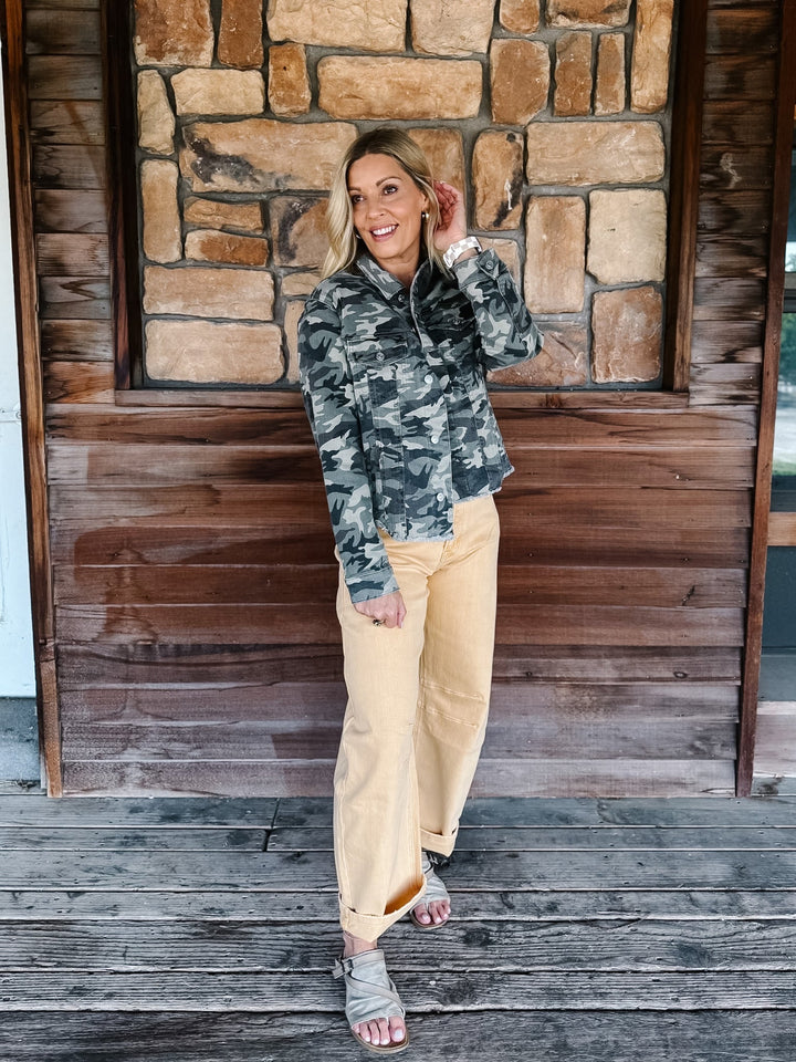 A woman stands on a rustic wooden porch in front of a stone wall, wearing a camouflage quarter-zip pullover jacket and light tan wide-leg pants.