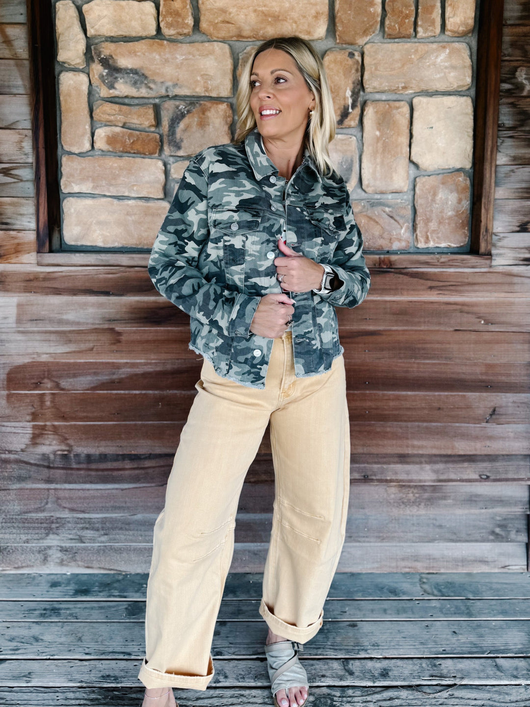A woman stands on a rustic wooden porch in front of a stone wall, wearing a green and gray camouflage button-up shirt and light beige wide-leg pants.