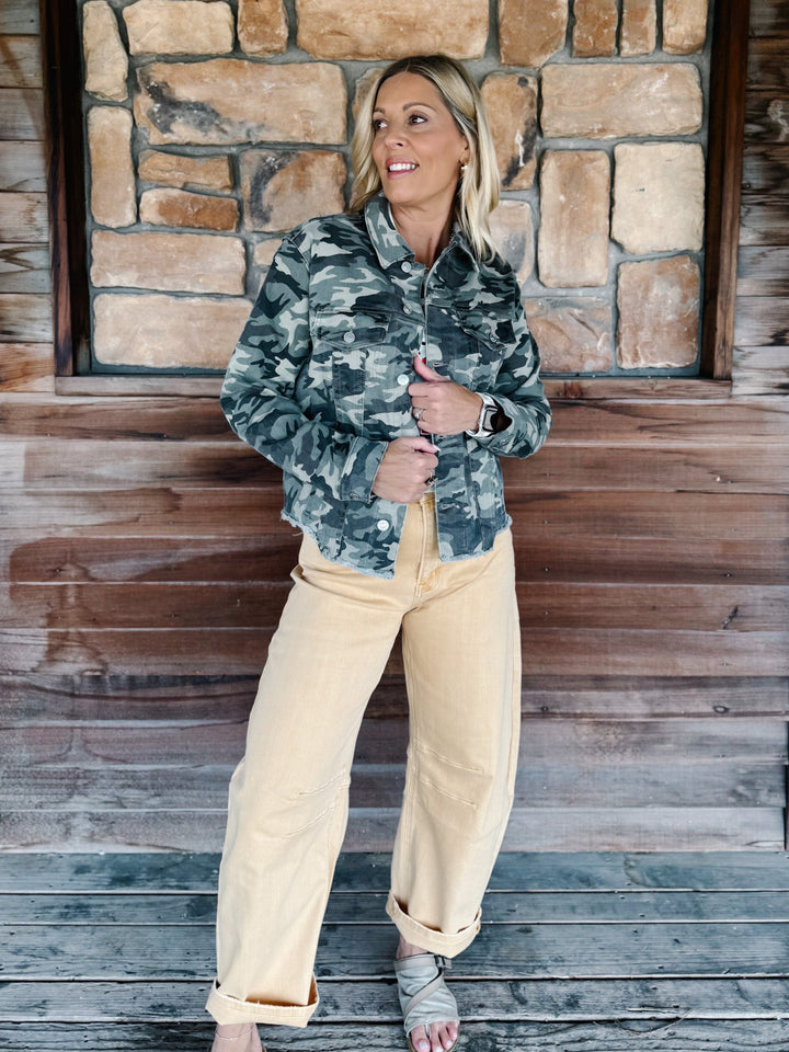 A woman stands on a rustic wooden porch in front of a stone wall, wearing a green and gray camouflage button-up shirt and light beige wide-leg pants.