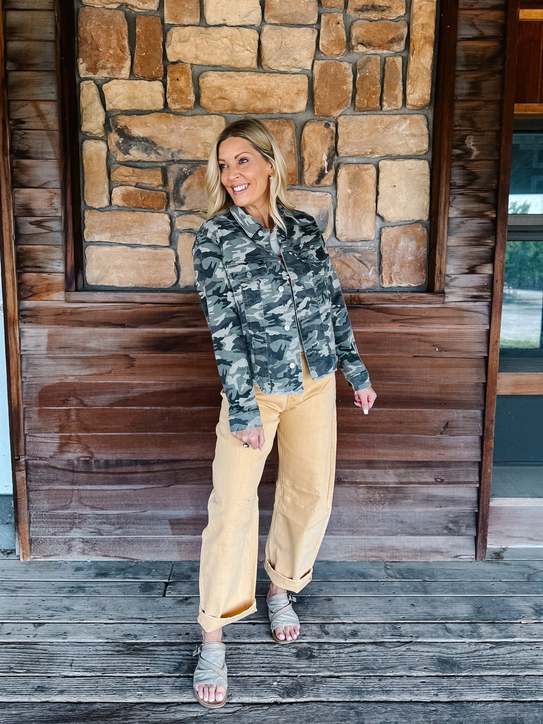 A woman stands on a wooden porch in front of a stone and wood wall, wearing a camouflage button-up shirt, tan wide-leg pants, and gray heeled sandals.
