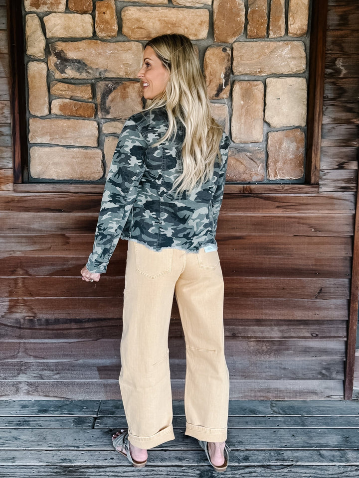 A woman stands on a rustic wooden porch in front of a stone wall, wearing a dark gray and black camouflage long-sleeve top and light beige wide-leg pants.