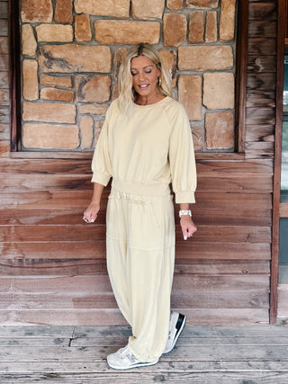 A woman wearing a light yellow-colored lounge set consisting of a long-sleeve sweatshirt and matching wide-leg pants, standing in front of a rustic stone wall and wooden floor.