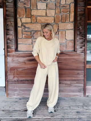 A woman wearing a light yellow-colored lounge set consisting of a long-sleeve sweatshirt and matching wide-leg pants, standing in front of a rustic stone wall and wooden floor.