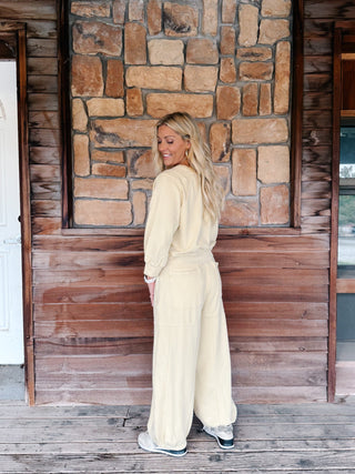A woman wearing a light yellow-colored lounge set consisting of a long-sleeve sweatshirt and matching wide-leg pants, standing in front of a rustic stone wall and wooden floor.