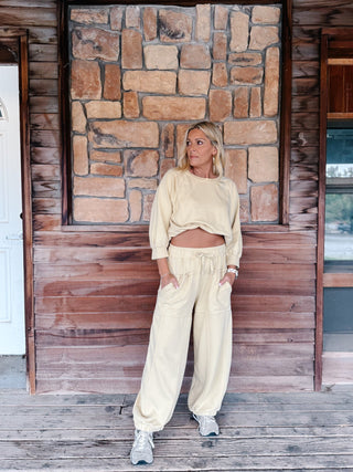 A woman stands on a wooden porch wearing a matching chamomile-colored lounge set consisting of a cropped long-sleeve top and wide-leg sweatpants, paired with gray sneakers.