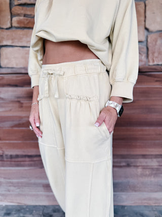 A woman stands on a wooden porch wearing a matching chamomile-colored lounge set consisting of a cropped long-sleeve top and wide-leg sweatpants, paired with gray sneakers.