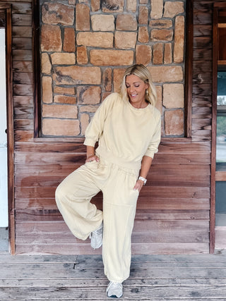 A woman stands on a wooden porch wearing a matching chamomile-colored lounge set consisting of a cropped long-sleeve top and wide-leg sweatpants, paired with gray sneakers.