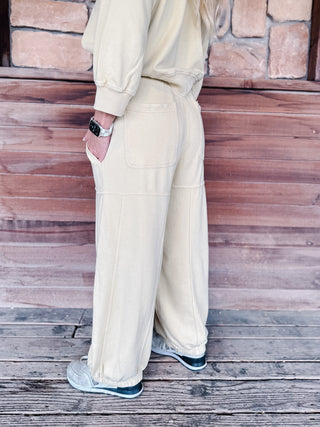 A woman stands on a wooden porch wearing a matching chamomile-colored lounge set consisting of a cropped long-sleeve top and wide-leg sweatpants, paired with gray sneakers.
