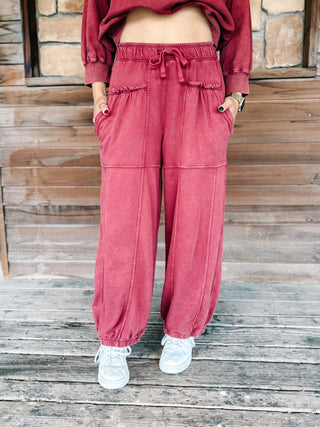 A person wearing crimson pants with a drawstring waist and elastic cuffs, paired with a matching sweatshirt and white sneakers, standing on a wooden deck.