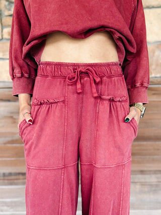 A person wearing crimson pants with a drawstring waist and elastic cuffs, paired with a matching sweatshirt and white sneakers, standing on a wooden deck.