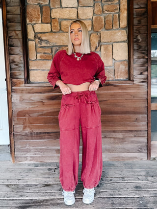 A person wearing crimson pants with a drawstring waist and elastic cuffs, paired with a matching sweatshirt and white sneakers, standing on a wooden deck.