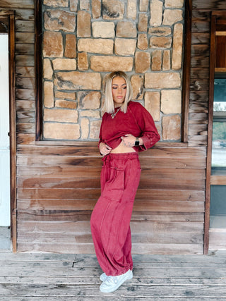 A person wearing crimson pants with a drawstring waist and elastic cuffs, paired with a matching sweatshirt and white sneakers, standing on a wooden deck.