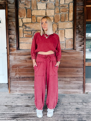 A person wearing crimson pants with a drawstring waist and elastic cuffs, paired with a matching sweatshirt and white sneakers, standing on a wooden deck.