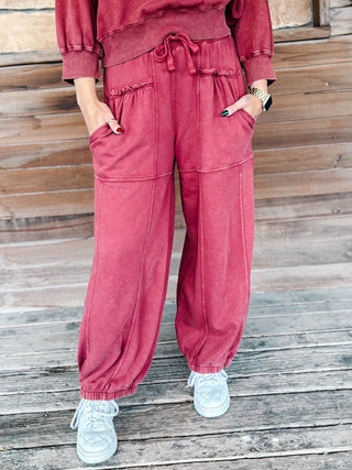 A person wearing crimson pants with a drawstring waist and elastic cuffs, paired with a matching sweatshirt and white sneakers, standing on a wooden deck.