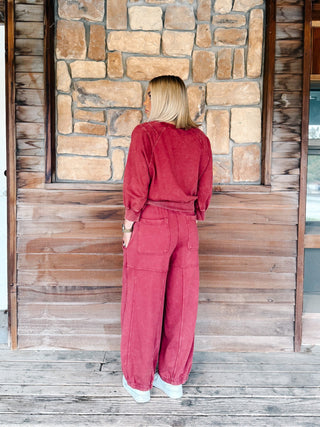 A person wearing crimson pants with a drawstring waist and elastic cuffs, paired with a matching sweatshirt and white sneakers, standing on a wooden deck.