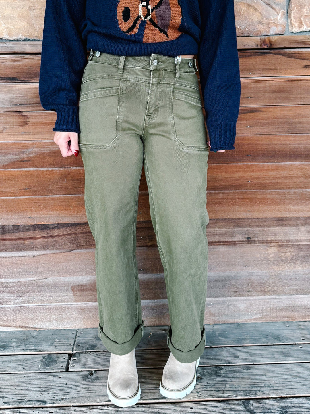 A person wearing olive green straight-leg jeans with rolled cuffs, paired with a navy sweater and beige boots, standing against a rustic wooden plank wall.