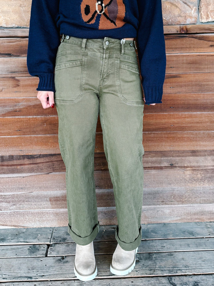 A person wearing olive green straight-leg jeans with rolled cuffs, paired with a navy sweater and beige boots, standing against a rustic wooden plank wall.