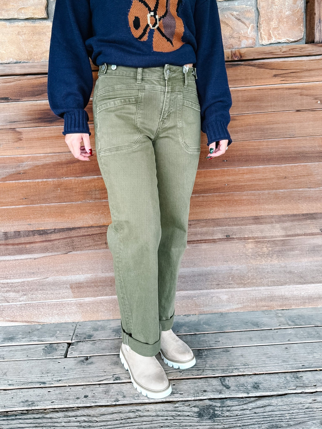 A person wearing olive green straight-leg jeans, a navy blue sweater with a brown graphic, and beige boots, standing against a rustic wooden plank wall.