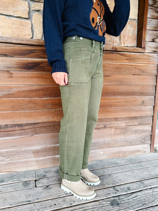 A person wearing olive green cargo-style pants, a navy sweater with a brown graphic, and beige boots, standing against a rustic wooden wall and deck.