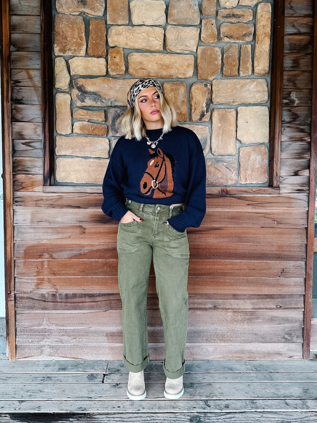 A woman stands on a rustic wooden porch in front of a stone wall, wearing a navy blue crewneck sweatshirt with a brown horse graphic, olive green jeans, and beige boots.