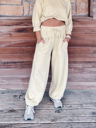 A person wearing camomile-colored lounge pants with a drawstring waist and side pockets, paired with a matching cropped sweatshirt and white sneakers, standing against a rustic wooden wall.