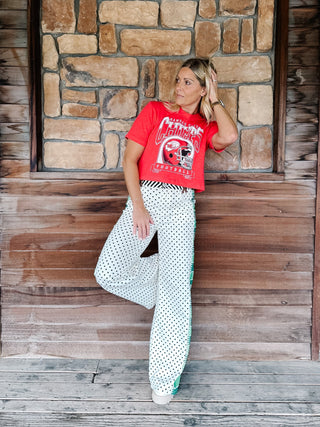 A woman posing in a red graphic t-shirt and white polka dot pants against a rustic wooden and stone wall background.
