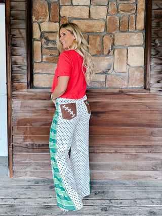 A woman stands on a wooden porch wearing a bright red short-sleeve t-shirt and wide-leg patchwork pants with a white and black polka dot pattern, green striped panels, and a brown football-shaped back pocket.