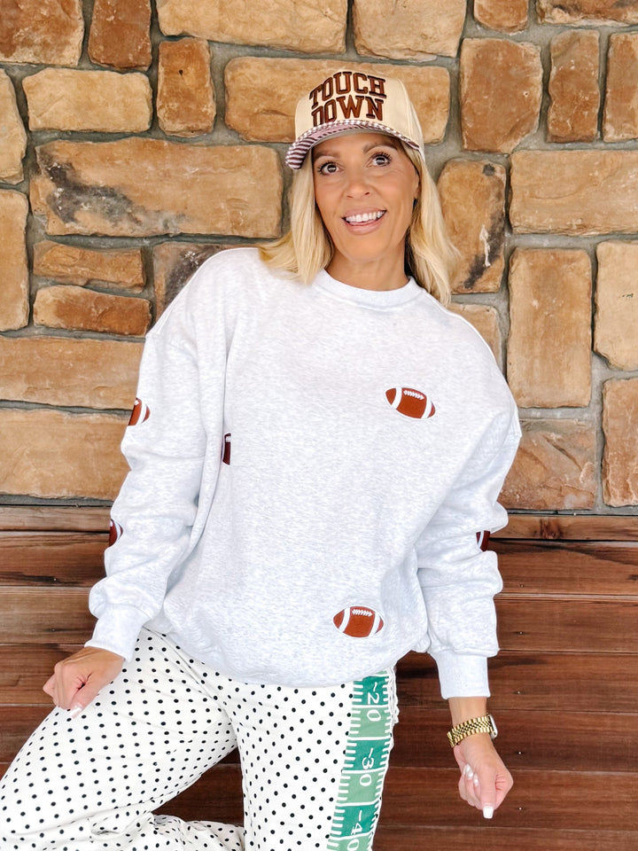A woman wearing a white heather crewneck sweatshirt with football graphics, white polka dot pants, and a beige cap that reads &