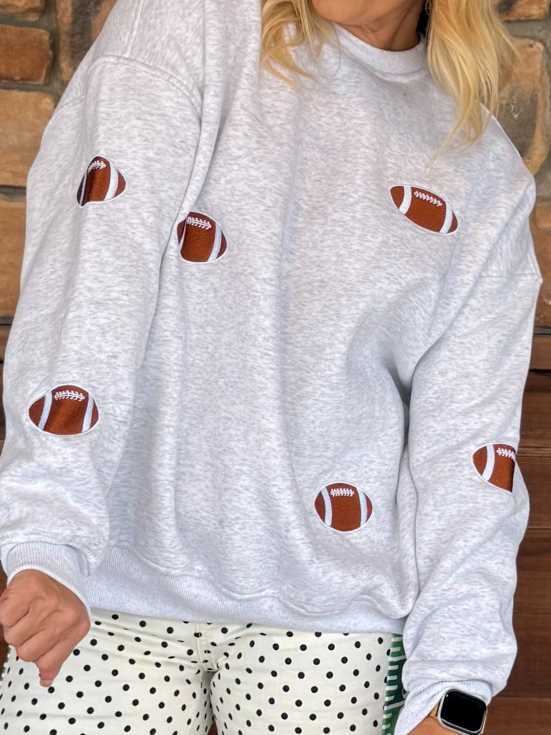 A person wearing a light heather gray sweatshirt with brown and white football patches sewn onto the front, paired with white pants with black polka dots, standing against a brick wall.