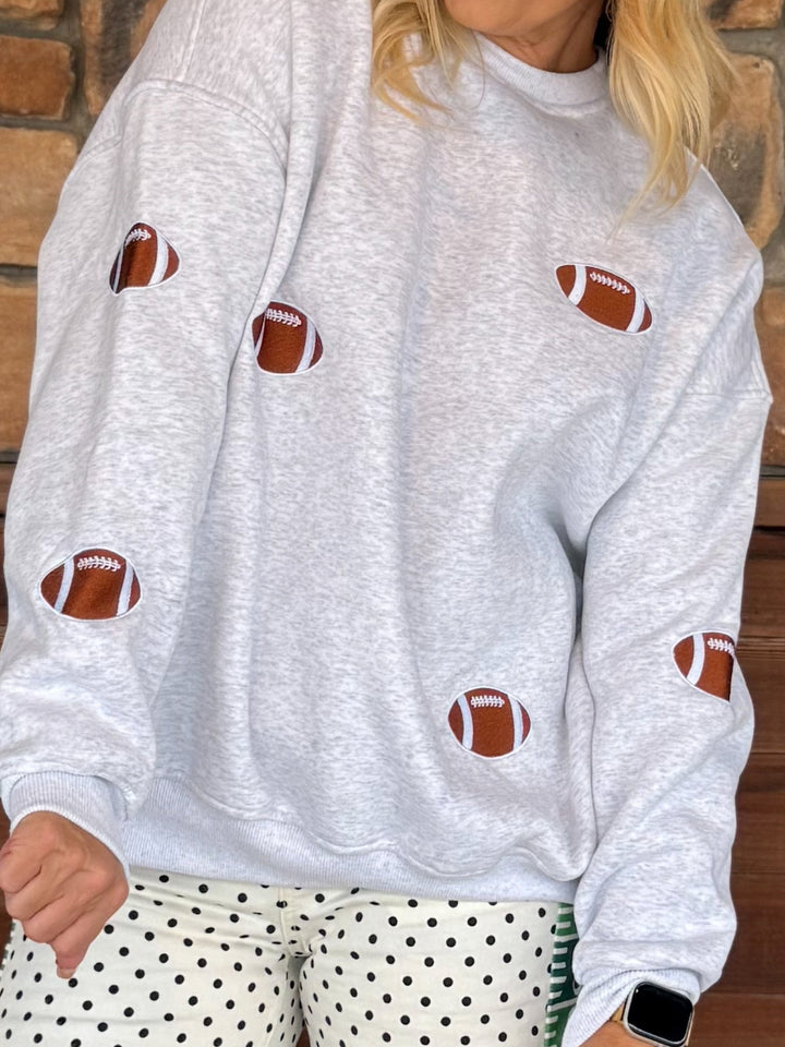 A person wearing a light heather gray sweatshirt with brown and white football patches sewn onto the front, paired with white pants with black polka dots, standing against a brick wall.